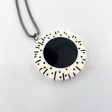 black and white resin circular pendant hung on oxidised silver chunky snake chain. The round pendant is cast in white resin and inlaid with a large black centre. The white edge is decorated with black lines and polka dots.