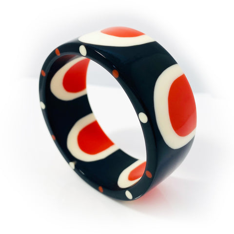 A shot of the beautiful abstract pop cuff standing upright. Cast in orange, white and black resin, it is inlaid with a sixties-inspired pattern and the edge is ornate with orange and white polka dots.