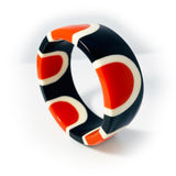 Shot of the abstract pop cuff standing upright. Cast in orange, white and black resin, it is inlaid with a sixties-inspired pattern.