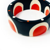 Close-up shot of the beautiful abstract pop cuff standing upright. Cast in orange, white and black resin, it is inlaid with a sixties-inspired pattern and the edge is ornate with orange and white polka dots.