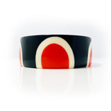 Upfront shot of the striking abstract pop cuff standing upright. Cast in orange, white and black resin, it is inlaid with a sixties-inspired pattern and the edge is ornate with orange and white polka dots.