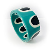 Flat surfaced large bangle inlaid with a sixties inspired pattern. Cast in a combination of black, white and teal blue resin. The bangle is upright, showing the edge ornate with black and white polka dots.