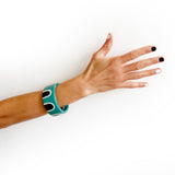 A wide patterned bangle worn on model's arm. The flat surfaced cuff is inlaid with a sixties inspired pattern. Cast in a combination of black, white and teal blue resin.