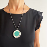 Large circle resin pendant cast with a teal centre and a white border, which is inlaid with black stripes. Hung on oxidised silver chunky snake chain. Worn on model.