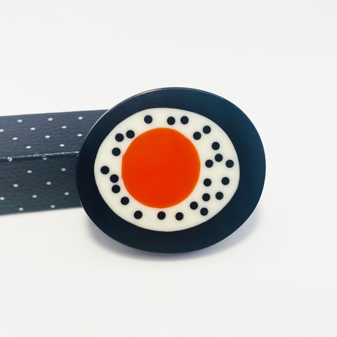 Oval abstract brooch handmade from resin. The centre is an orange circle, which is bordered with white resin,  which is itself encircled with black resin. The white band is inlaid with scattered polka dots.
