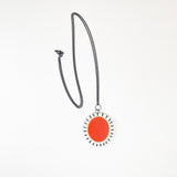 Large oval shaped pendant cast with a red centre and a white border which is inlaid with black stripes. It is hung on a long oxidised silver chain.