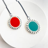 2 large oval shaped resin pendants photographed on a white circle display. One has a red centre, the other has a teal centre. Both are cast with a cream border which is inlaid with black stripes. They are hung on oxidised silver chain.