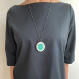 Large oval shaped teal blue and white long pendant worn by model on a dark grey dress.