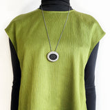 Person wearing a green sleeveless top with a black turtleneck underneath, displaying a long necklace with a circular pendant.