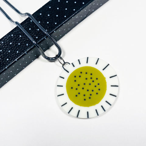 Oval-shaped large resin pendant cast with an olive green middle, inlaid with black polka dots. Its white border is inlaid with black stripes. Hung on chunky silver snake chain.