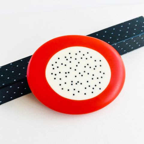 Pebble shaped oversized resin brooch cast with a cream centre and an orange border. The centre is inlaid with small black dots. The brooch is displayed against a polka and white dotted box.