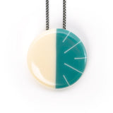 The Lunula circular pendant is a beautiful modernist statement piece, handmade from resin and Sterling silver. One half is cast in cream, the other is teal blue, ornate with contrasting lines in white. It hangs on a 70cm Sterling silver oxidised chain. 